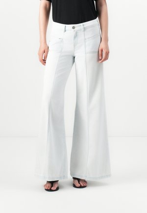 Femme portant un pantalon blanc taille haute, jambes larges avec des coutures devant, assorti à un haut noir et des sandales noires à bouts ouverts.