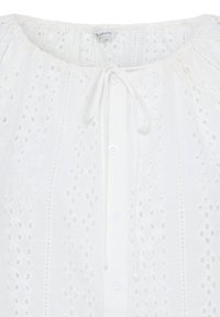 Blouse blanche en dentelle à œillets avec col rond, boutons sur le devant et un détail de ruban au col, mettant en valeur un motif brodé délicat.