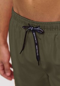 Detail osoby nosící olivově zelené šortky s černým šňůrkovým páskem uvázaným na mašli, s nápisem „U.S. POLO ASSN.“ na šňůrce.