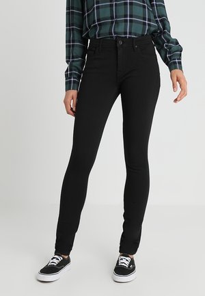 Personne portant un jean skinny noir, une chemise à carreaux verte rentrée dans le pantalon, et des baskets noires avec des semelles blanches, debout sur un fond clair uni.