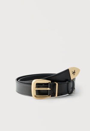 Cinturón de cuero negro enrollado con hebilla en tono dorado y punta metálica grabada con el logo "ck", colocado sobre un fondo liso y claro.