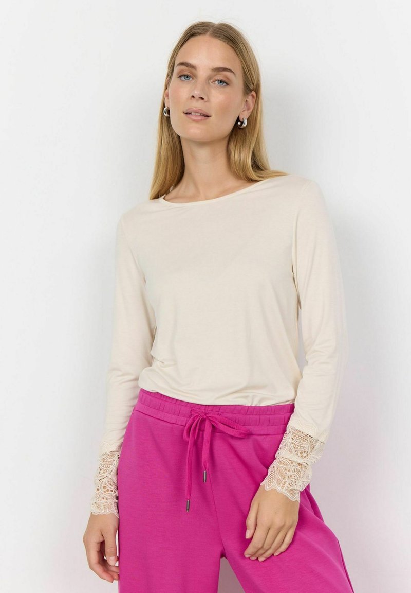 Beige långärmad topp med spetsdetaljer vid ärmsluten, i kombination med rosa byxor med dragsko. Mjuk textur och avslappnad passform.