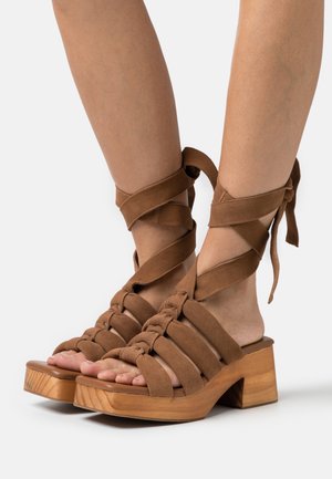 Sandales à talons en daim marron avec un design tissé et des liens à la cheville. Talons épais en bois, semelle lisse, bout ouvert et texture subtile.