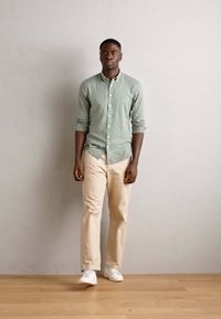 Cămașă button-up de in verde deschis, pantaloni bej și adidași albi. Modelul stă în fața unui perete neutru, cu un podea din lemn vizibilă.