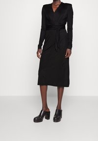 Schwarzes Wickelkleid mit langen Ärmeln, V-Ausschnitt und Taillenband. Glatter Stoff in Midi-Länge. Kombiniert mit schwarzen Blockabsatz-Mules.