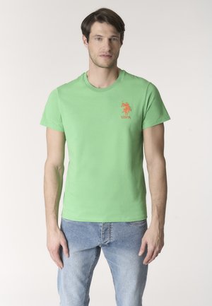 Uomo che indossa una t-shirt verde chiaro con il logo USPA e jeans azzurri, in piedi contro uno sfondo chiaro uniforme.