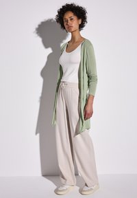 Lichtgroene lange cardigan over een witte tanktop, gecombineerd met beige wijde pantalon en lichte sneakers. Zachte textuur, casual stijl.