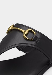 Μαύρο δερμάτινο σανδάλι σλάιντ με χρυσή μεταλλική λεπτομέρεια σε σχήμα αλογόμυγας στη λουρίδα στο πάνω μέρος, πάνω σε λευκό φόντο.