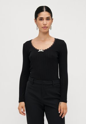 Haut noir à manches longues avec une texture côtelée, décolleté bordé de dentelle et un nœud décoratif blanc au centre. Porté avec un pantalon noir.