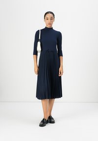 Navy ribbed midi dress with short sleeves, pleated skirt, and drawstring waist. Paired with black shoes and a small white handbag.