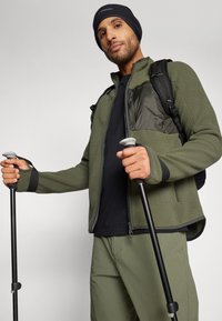 Giacca in pile verde, dettagli neri e tasche con zip, indossata con un berretto nero. Tieni in mano dei bastoni da trekking; pantaloni khaki completano l'outfit.
