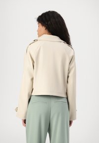 Beige kort jacka med krage och raglanärmar, med en mjuk textur. Bärs över ljusgröna skräddarsydda byxor.