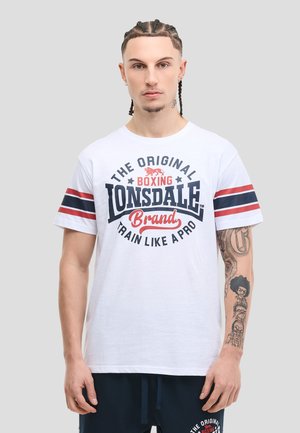 Mężczyzna z plecionymi włosami w białej koszulce bokserskiej Lonsdale z czerwonymi i granatowymi paskami oraz ciemnymi szortami, stojący na szarym tle.