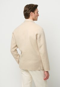 Giacca in misto lana beige con design su misura, spacco singolo sul retro e maniche lunghe. Texture liscia, dettagli minimi e linee pulite.