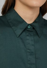 Chemise col montant vert foncé avec un bouton de fermeture, texture lisse et col structuré avec des pointes nettes.