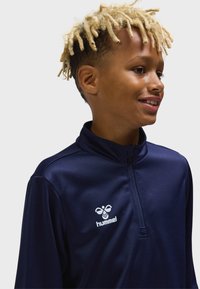 Marinblå atletisk tröja med kvartslång dragkedja i slätt, lätt material. Har en vit logga på bröstet och en stående krage.