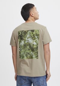 Khaki tričko s velkým zeleným grafikou tropického listu na zadní straně. Měkká látka s kulatým výstřihem a krátkými rukávy.