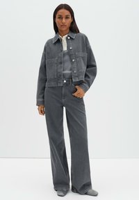 Vrouw met een grijs spijkerjack met grote voorzakken, bijpassende wijde jeans, een wit overhemd met kraag, en grijze puntige schoenen.
