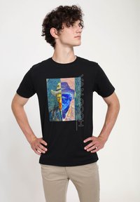 Mladý muž s kudrnatými vlasy, oblečený v černém tričku s rozděleným barevným van Goghovým autoportrétem a textem "Van Gogh," stojící s rukama v bok.