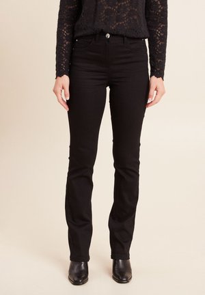 Frau trägt ein langärmeliges Oberteil aus schwarzer Spitze, hochtaillierte schwarze Slim-Fit-Jeans und schwarze Stiefeletten, steht vor einem einfarbigen Hintergrund.