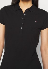 Černé polo tričko s límečkem, krátkými rukávy a légou na knoflíky se stříbrnými knoflíky. Na hrudi je malé vyšívané logo.