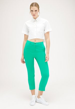Witte cropped shirt met een kraag en knopen aan de voorkant, gecombineerd met felgroene fitted broek en witte sneakers, staande tegen een effen achtergrond.