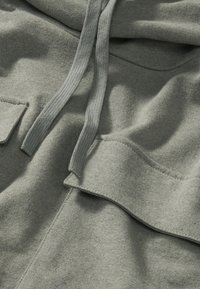 Felpa verde chiaro con un tessuto texturizzato, tasca frontale con flap e cordoncini. Il materiale sembra morbido e i bordi sono rifiniti.