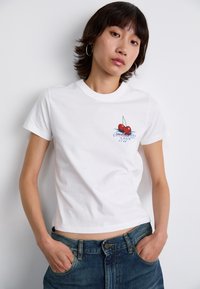 Camiseta de algodón blanca con un gráfico de cereza y un diseño de texto en el lado izquierdo, combinada con unos vaqueros de mezclilla azul. Mangas cortas, cuello redondo.