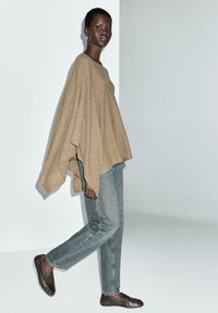 Poncho khaki au texture lisse et aux manches larges, associé à un jean droit bleu clair et des chaussures à enfiler marron foncé.