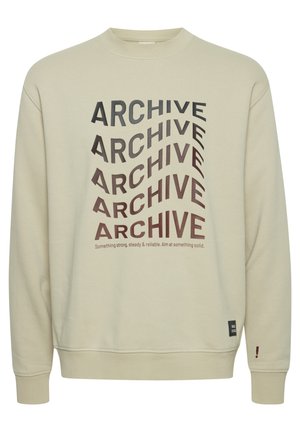 Beež müts, mis on valmistatud puuvillast, esitleb julgeid musti ja pruunikaid "ARCHIVE" tekste erinevates suundades, ribatud varrukaid ja sildi aktsenti.