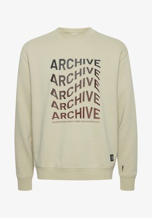Beige genser laget av bomull, med dristig svart og brun "ARCHIVE" tekst i varierende retninger, ribbede mansjetter og en etikett som aksent.