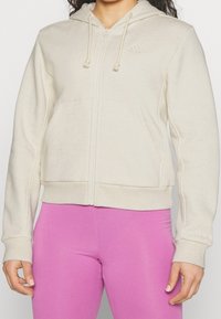 Osoba v krémovém zipovém svetru s předními kapsami a růžovými legínami, zachycena od ramen po boky, stojící s uvolněnýma rukama.