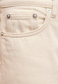 Μπεζ ύφασμα τζιν σε κοντινό πλάνο που δείχνει μπροστινή τσέπη με μεταλλικά καρφιά και λεπτομερείς ραφές στο παντελόνι.