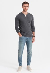 Mann steht mit den Händen in den Taschen, trägt ein dunkelgraues Langarm-Henley-Shirt, hellblaue Jeans, weiße Sneakers und ein schwarzes Armband.