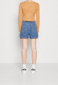 Jeansshorts med hög midja, medelblå tvättning och en ren fåll. Parat med en långärmad topp i orange. Vita strumpor och svarta sneakers.