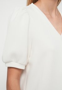 Λευκό πουκάμισο με V-λαιμόκοψη, φουσκωτά κοντά μανίκια και απαλό ύφασμα. Ο σχεδιασμός είναι μινιμαλιστικός χωρίς ορατά σχέδια ή διακοσμητικά στοιχεία.