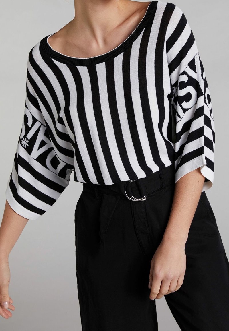 Personne portant un haut à rayures verticales noires et blanches avec un col bateau et des manches mi-longues, associé à un pantalon noir taille haute et une ceinture à double boucle.