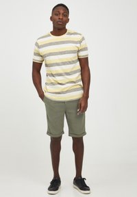 Kortermet stripet t-skjorte i gul, grå og hvit, kombinert med olivengrønne shorts og svarte sneakers, stående på en nøytral bakgrunn.
