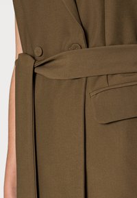 Brun tyll coat med bälte i midjan, två runda knappar och en sidficka med vikbar flikdetalj. Slät textur, skräddarsydd design.