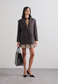 Ruskea bleiseri, jossa on kaksi nappia, beige paita, jossa on napit, ruudullinen hame, mustat korkokengät ja musta käsilaukku. Yhtenäiset sileät tekstuurit.