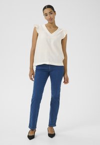 Blouse crème à col en V avec des manches à volants, associée à un jean bleu et des escarpins noirs à bout pointu. Tissu lisse, coupe décontractée et design à jambe droite.