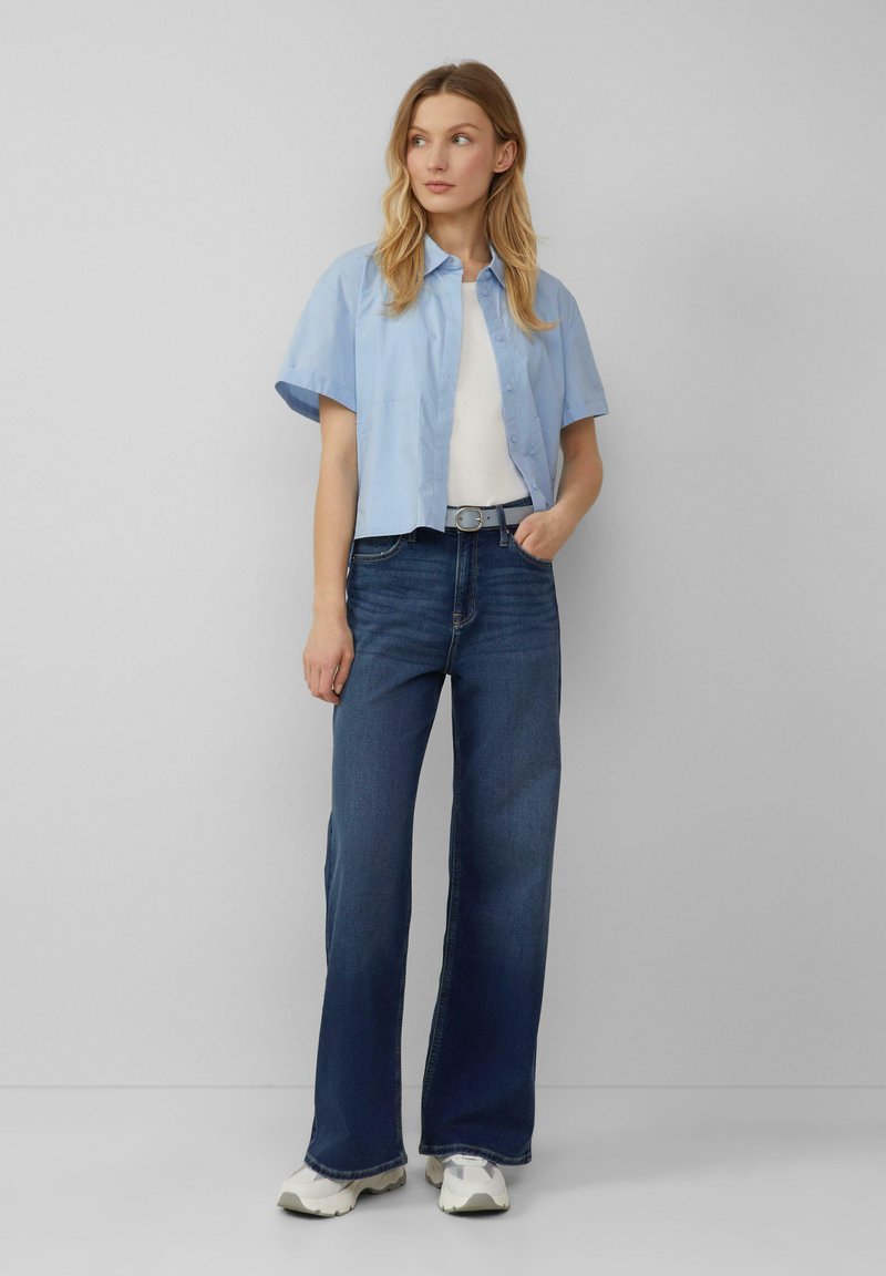 Lichtblauwe short-sleeve shirt, witte top eronder, gecombineerd met hooggesneden donkerblauwe wijde jeans. Witte sneakers, casual look.