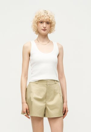 Femme blonde aux cheveux bouclés portant un haut blanc sans manches, un short beige taille haute et un fin collier en or, debout devant un fond uni.
