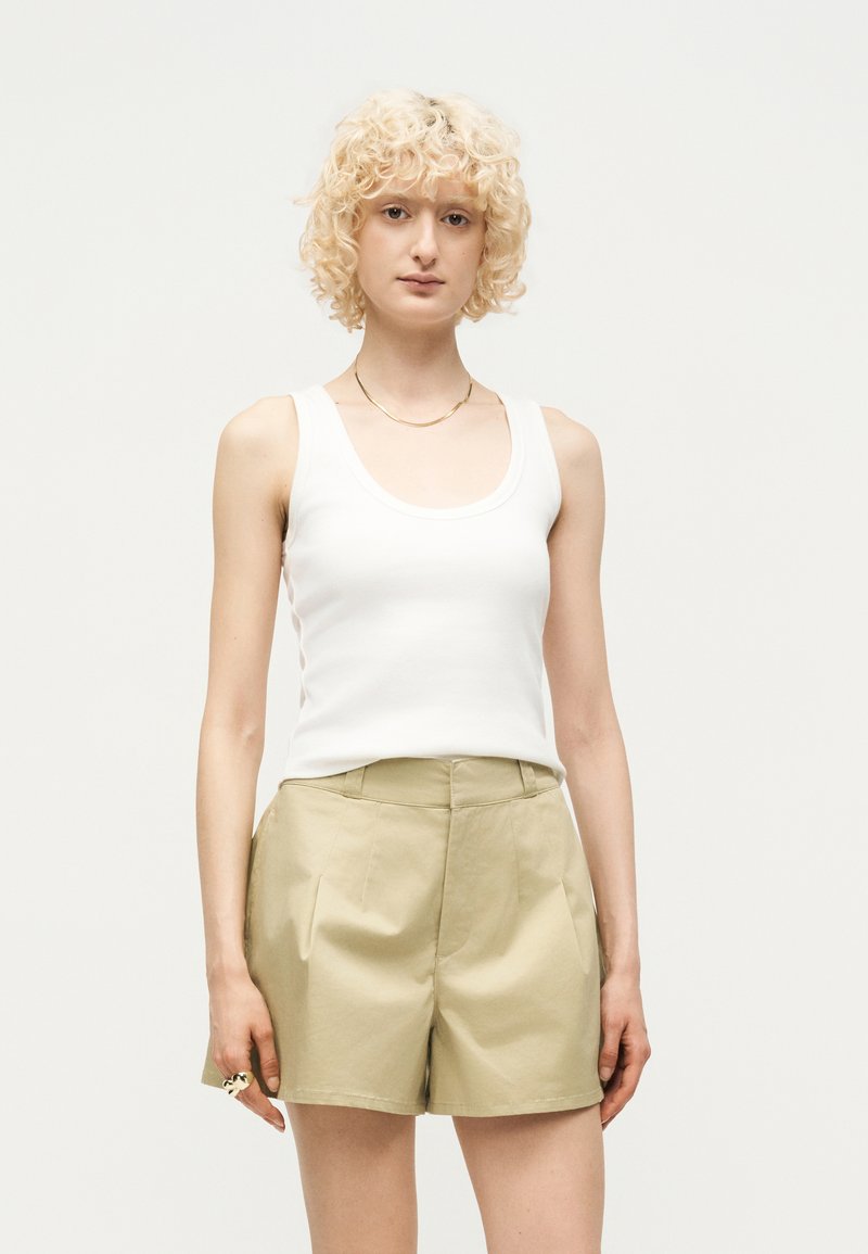 Ξανθιά γυναίκα με κατσαρά μαλλιά, που φοράει λευκό αμάνικο τοπ, μπεζ σορτς με ψηλή μέση και λεπτό χρυσό κολιέ, στέκεται μπροστά σε μια απλή φόντο.