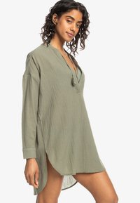 Abito tunica a maniche lunghe, di colore verde oliva, realizzato in tessuto leggero. Presenta un scollo a V, spacchi laterali e una vestibilità rilassata. Superficie testurizzata e dettaglio del colletto.