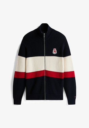 Gebreide ritscardigan in marineblauw, crème en rode strepen. Beschikt over een hoge kraag en een merkembleem aan de linkerkant. Geribbelde textuur.