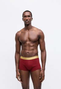 Röda boxershorts med ett strukturerat guldmidjeband, med en nära passform och slät tyg, visade på en modell mot en enfärgad bakgrund.