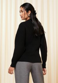 Schwarzer, gerippter Strickpullover mit langen Ärmeln, tailliert an der Taille. Kombiniert mit grauen, maßgeschneiderten Hosen vor einem hellbeigen Hintergrund.