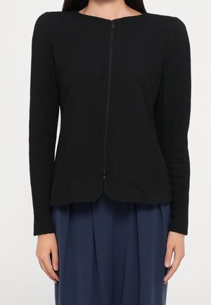 Femme portant une veste noire ajustée texturée à manches longues avec fermeture éclair devant, sur un pantalon bleu marine ample.