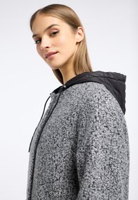 Grijze, textuur-achtige hoodie met een zwarte gewatteerde capuchon. Voorzien van een rits en verstelbare trekkoorden. Zacht en comfortabel materiaal.